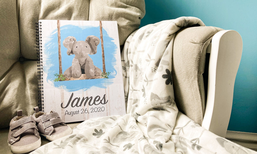 Elephant baby book sitting on grey cushioned rocking chair alongside white baby blanket and grey baby shoes